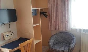 A simple room with a desk, a chair, and a shelf. A television hangs on the wall, and the window lets in a lot of daylight.