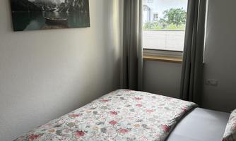 A cozy bedroom with a double bed and floral bedding. The room has a window with curtains and a modern light fixture.