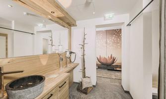 A modern bathroom with wooden paneling and an elegant sink. In the background, there is a relaxing wellness area decorated with a lotus flower.