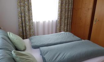 A cozy bedroom with a double bed and fresh pillows. The windows are decorated with curtains and there is a wooden wardrobe in the room.