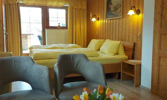 A cozy room with wooden walls and a large window. There is a double bed, two chairs, and a table with a vase full of flowers.