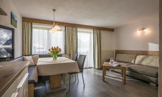 A modern living room with a table, chairs, and a cozy couch. Large windows let in a lot of light and there are fresh flowers on the table.
