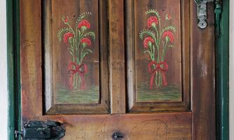 An old wooden door with colorful flower motifs and simple metal fittings. Above are the years 18, C, M, B, and 72 engraved in white.