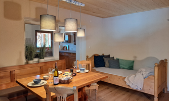 A cozy dining area with a wooden chair and a table set for meals. Against the wall stands a comfortable lounger, surrounded by warm light and natural wood paneling.
