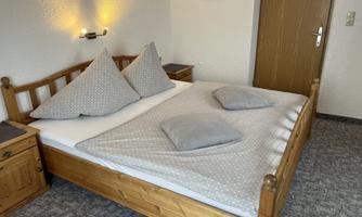 A cozy bedroom with a wooden bed and two decorative pillows. The walls are light and there is warm lighting.