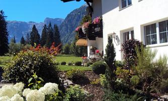 A beautiful house with blossoming balcony plants and a well-maintained garden. In the background, tall mountains and a clear blue sky can be seen.