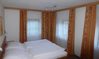 A bright bedroom with a double bed and white bedspreads. The walls are designed with wooden paneling and windows with curtains.