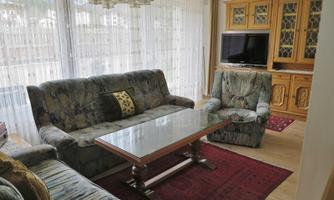 A cozy living room with a large sofa, an armchair, and a wooden table. The windows are adorned with light curtains and a television stands in the corner.