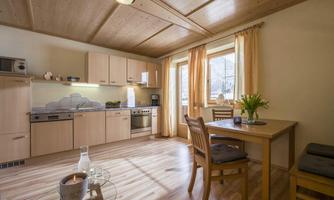 A modern kitchen with wooden cabinets and a dining area. The room has large windows and is bright and friendly.
