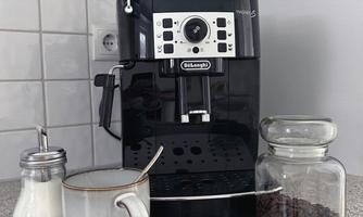 A modern coffee machine stands on a countertop. Next to it are a cup, sugar, and a glass with coffee beans.