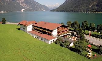 A picturesque landscape with a large lake and mountains in the background. In the foreground stands an attractive building surrounded by lush greenery.