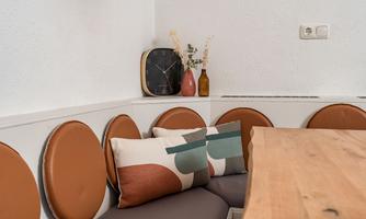 A cozy dining area with a wooden table top and an elegant corner bench. Decorative cushions, a clock, and vases add a stylish touch to the room.