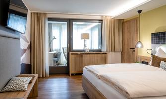 A modern hotel room with a double bed and wooden furniture. Large windows provide plenty of daylight and offer a view of the outside.