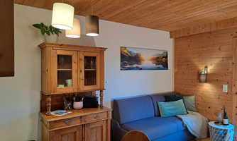 A cozy living room with a blue sofa and a wooden display cabinet. On the wall hangs a picture of a landscape with water and mountains.