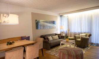 A modern furnished living room with a cozy sofa and an armchair. A dining table with chairs and large windows provide plenty of light.