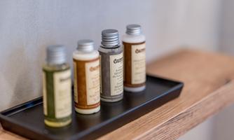 Four small bottles of shampoo and shower gel are standing on a black tray. The background is simple and modern.