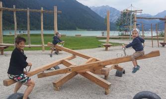 A woman and two children are playing on a wooden swing at a playground. In the background, mountains and a lake can be seen.