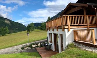 A charming house with a wooden terrace in a quiet, green landscape. In the background, there are hills and a blue sky.