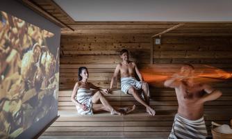 The image shows three people in a sauna. One person is conducting a ritual with a splash of water, while the others are relaxing.