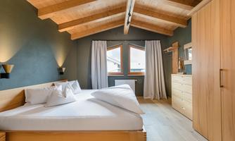 A modern bedroom with a wooden ceiling and large windows. It is cozily furnished with a double bed and a wooden wardrobe.
