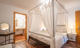 A cozy bedroom with a canopy bed and white curtains. In the background, a modern bathroom can be seen.