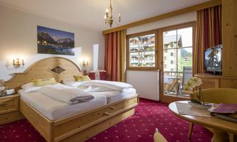 A cozy hotel room with a large bed and traditional wooden decor. Large windows offer a view outside, and the room is furnished with warm colors and modern furniture.