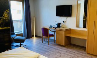 A modern hotel room with a double bed, a desk, and a television. Large windows bring plenty of daylight into the room.