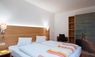 A modern bedroom with two single beds and light wooden furniture. The room includes a desk and a wardrobe with a decorative picture.