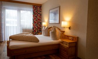 A cozy bedroom with a double bed, two pillows, and wooden furniture. Daylight streams through large windows, creating an inviting atmosphere.