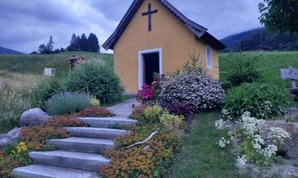 A small yellow chapel with a cross on the roof. Colorful flowers bloom around the chapel, and the garden is well-maintained.