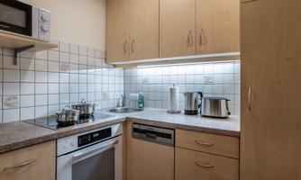 A modern kitchen with wooden furniture and white tiles. It is equipped with a stove, an oven, and various kitchen appliances.