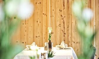 An elegant restaurant with a bright wooden wall and a beautifully set table. On the table, there is a flower in a vase, surrounded by minimalist decoration.