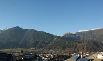 An impressive mountain landscape under a clear blue sky. In the foreground, some houses and roofs are visible.