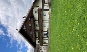 A large, multi-story house with a slanted roof. It is located in a green meadow under a blue sky with clouds.