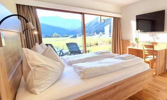 A modern room with a double bed and large windows. The view shows a picturesque mountain landscape.