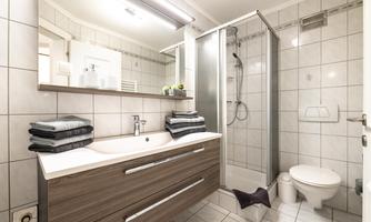 A modern bathroom with a glass shower, a wooden washbasin, and large, bright tiles. Dark grey towels are placed on the washbasin and a small plant is in the background.