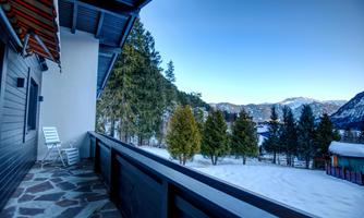 A balcony with a view of snowy mountains and coniferous trees. The sky is clear and blue.