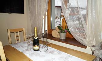 A table with a bottle of champagne, glasses, and candles. Decorative curtains and a plant pot are placed in front of a window.