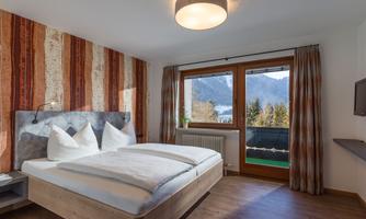 A modern bedroom with a double bed and wooden flooring. Large windows provide a view of the mountains and nature.