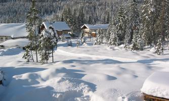 A snowy landscape with small huts and tall fir trees. The snow covers the entire surroundings and the sky is clear.