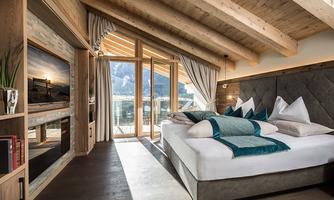 A modern bedroom with a wooden ceiling and large windows. It is tastefully furnished with cushions and a cozy bed.