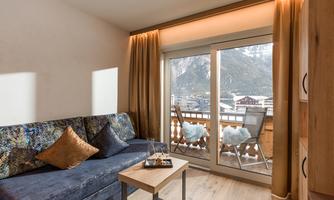 A modern living room corner with a cozy sofa and a wooden coffee table. Large windows offer a view of the mountains and the balcony.