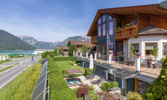 A cozy house with a balcony and garden, surrounded by a picturesque mountain landscape. The sun shines on the adjacent road and the nearby lake.