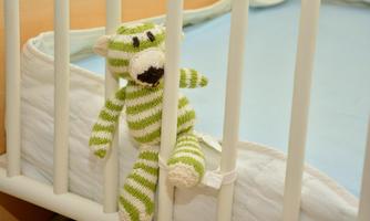 A knitted stuffed bear is sitting on the bars of a crib. The bear has green and white stripes and looks friendly.