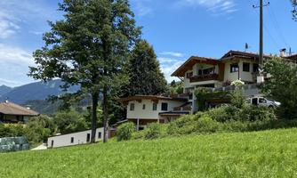 A picturesque house in a green landscape, surrounded by trees and mountains. The sky is clear and blue.