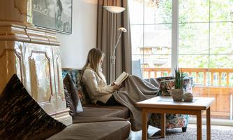A person is sitting comfortably on a sofa and reading a book. The room is bright and inviting with a view of nature through the window.