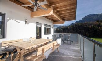 A spacious terrace with a wooden chair and a table. In the background, green mountains and a blue sky can be seen.