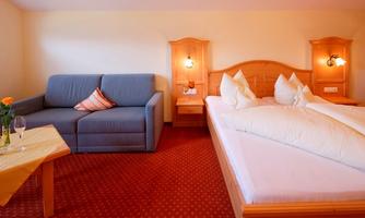 A cozy room with a double bed and a gray sofa. On the table, there is a flower and two glasses.