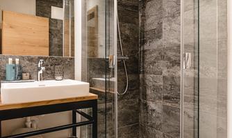 A modern bathroom with a glass shower and an elegant sink. The walls are designed in a dark stone look, and there are neatly folded towels on the shelf.