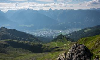 A breathtaking mountain landscape with green meadows and majestic peaks in the background. In the valley, a town is visible, surrounded by mountains and nature.